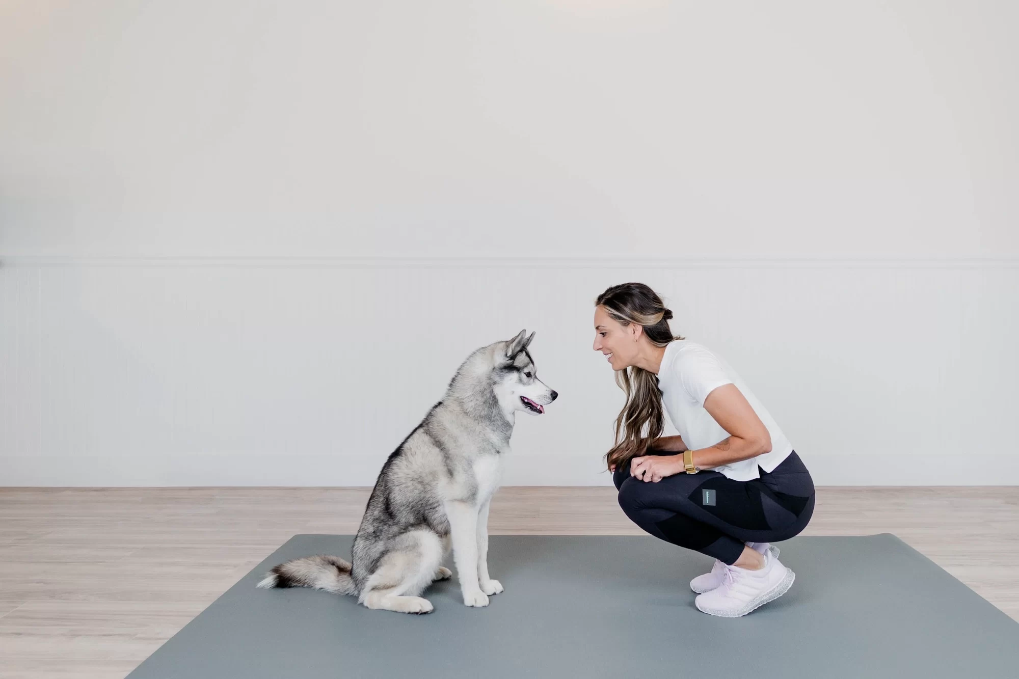 Woman in athletic wear crouching on exercise mat beside sitting husky dog in bright studio setting
